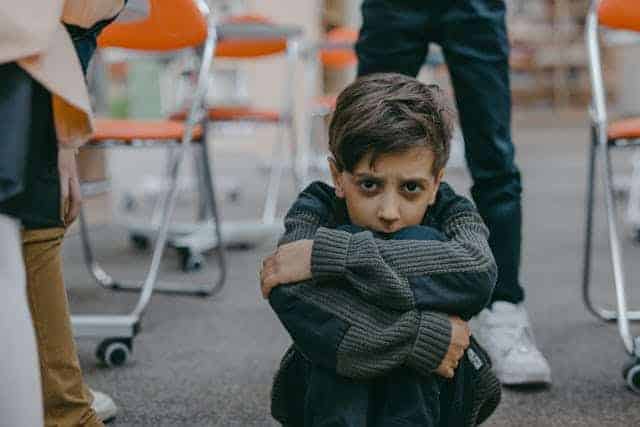 Hate boy wearing a gray sweater sits on the ground, arms around his knees, looking scared.