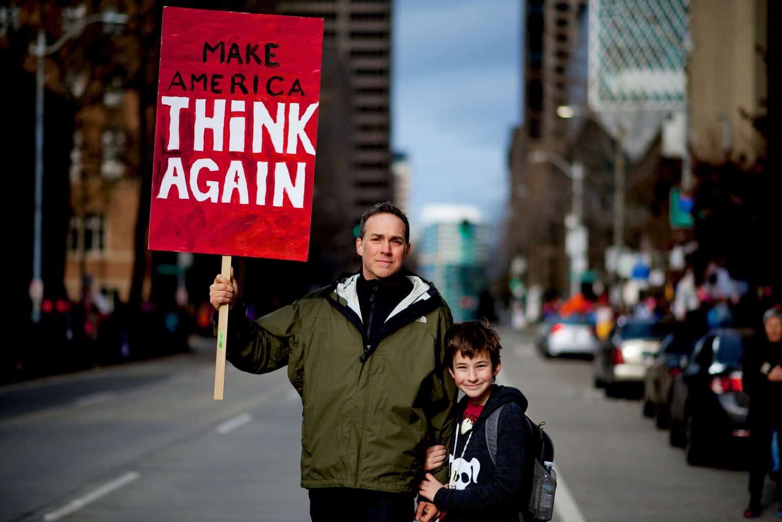 Father and son stand in the street of a city holding a sign that says make america think again