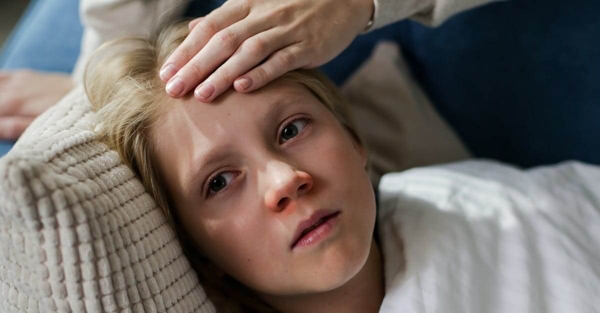 Mixed messages about measles from rfk jr young child lying down with a hand checking for fever, indicating illness or flu symptoms.