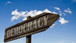 Wooden democracy sign with a blue sky in the background. What is in project 2025