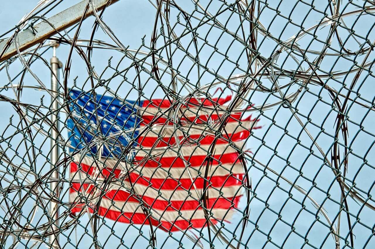 Razorwire and fencing in front of a torn american flag. 6 deaths in california ice detention